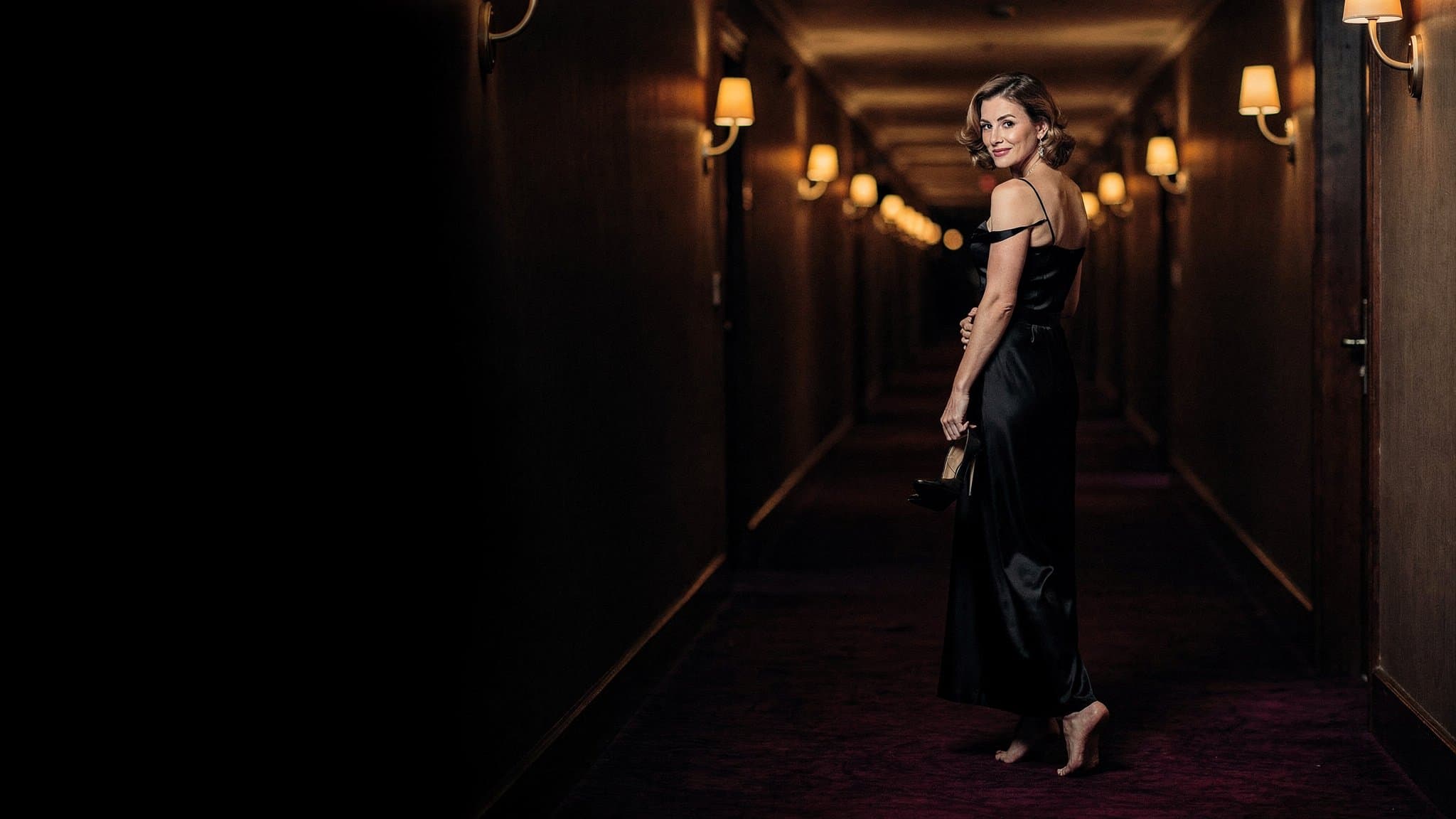 A woman in a black dress walking down a candlelit hotel corridor, glancing back at you